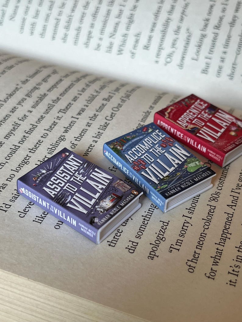 May include: Three miniature books with the titles "Assistant to the Villain," "Accomplice to the Villain," and "Apprentice to the Villain" are displayed on an open book. The books have colorful covers with illustrations and text. The pages of the open book are visible in the background.