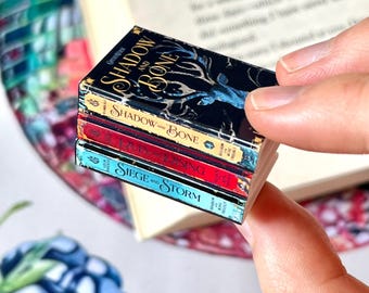 May include: Miniature book stack featuring the titles "Shadow and Bone", "Siege and Storm", and "Shadow and Bone". The books have colorful spines and detailed covers. The stack is held in a person's hand, with a larger book and patterned background visible.