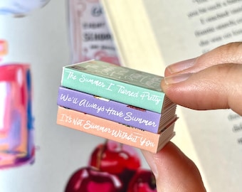 May include: A stack of miniature books with pastel covers and titles: "The Summer I Turned Pretty," "We'll Always Have Summer," and "It's Not Summer Without You." The books are held in front of a blurred background with cherries.