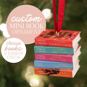 May include: A stack of five miniature book ornaments hanging from a red ribbon. The books are titled A Court of Thorns and Roses, A Court of Mist and Fury, A Court of Wings and Ruin, A Court of Frost and Starlight, and A Court of Silver Flames by Sarah J. Maas.