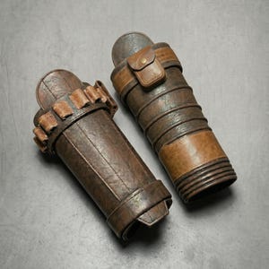 May include: Two brown leather quivers with varying shades and textures. One quiver has a series of small, rectangular holders, while the other features a small flap with a button closure. Both have a cylindrical shape and are designed to hold arrows.
