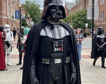 Könnte beinhalten: Ein Darth-Vader-Kostüm mit schwarzem Helm, langem schwarzem Umhang und einem Brustbedienfeld mit roten und blauen Knöpfen. Der Anzug umfasst schwarze Handschuhe, einen Gürtel mit silbernen Akzenten und einen strukturierten schwarzen Anzug.