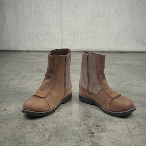 May include: A pair of brown ankle boots with a side zipper and a contrasting light brown panel. The boots have a rounded toe and a dark sole. The boots are made of a suede-like material.