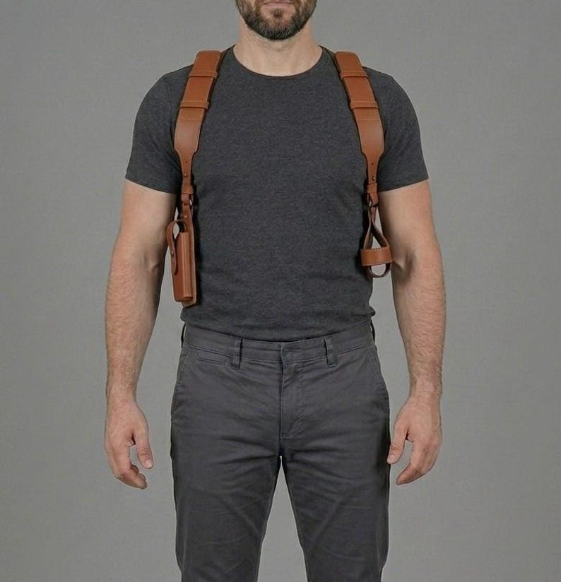 May include: A tan leather shoulder holster system featuring wide, adjustable straps that cross over the shoulders and connect to a vertical holster on the right side and a circular loop attachment on the left side, worn over a dark grey t-shirt.