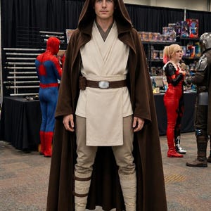 Puede incluir: Una persona con una capa con capucha marr&oacute;n y una t&uacute;nica, pantalones y botas beiges, que se asemeja a un Jedi de Star Wars. Un cintur&oacute;n marr&oacute;n con hebilla plateada y una bolsa completan el disfraz. Otras figuras disfrazadas est&aacute;n en el fondo.