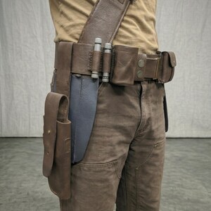 Puede incluir: Un cintur&oacute;n utilitario de cuero marr&oacute;n con m&uacute;ltiples bolsas, una funda y una correa para el hombro. El cintur&oacute;n presenta detalles cil&iacute;ndricos plateados y se usa con pantalones marrones. Un objeto azul es parcialmente visible.