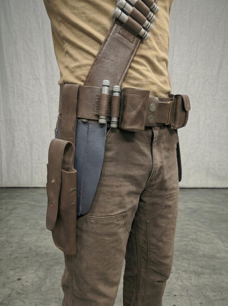 Puede incluir: Un cintur&oacute;n utilitario de cuero marr&oacute;n con m&uacute;ltiples bolsas, una funda y una correa para el hombro. El cintur&oacute;n presenta detalles cil&iacute;ndricos plateados y se usa con pantalones marrones. Un objeto azul es parcialmente visible.