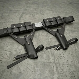 May include: A black utility belt featuring six small rectangular pouches with snap closures, a central metallic buckle, and two attached holsters. Each holster is secured with adjustable straps and metal buckles, creating a structured tactical design.