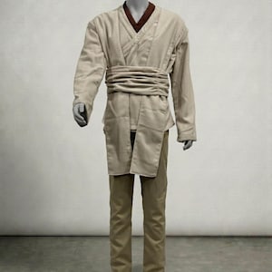 Puede incluir: Un disfraz de color beige compuesto por una t&uacute;nica de manga larga, pantalones y un cintur&oacute;n ancho. La t&uacute;nica tiene un cuello marr&oacute;n y un dise&ntilde;o envolvente. Los pantalones son de color beige a juego. El disfraz se exhibe en un maniqu&iacute;.