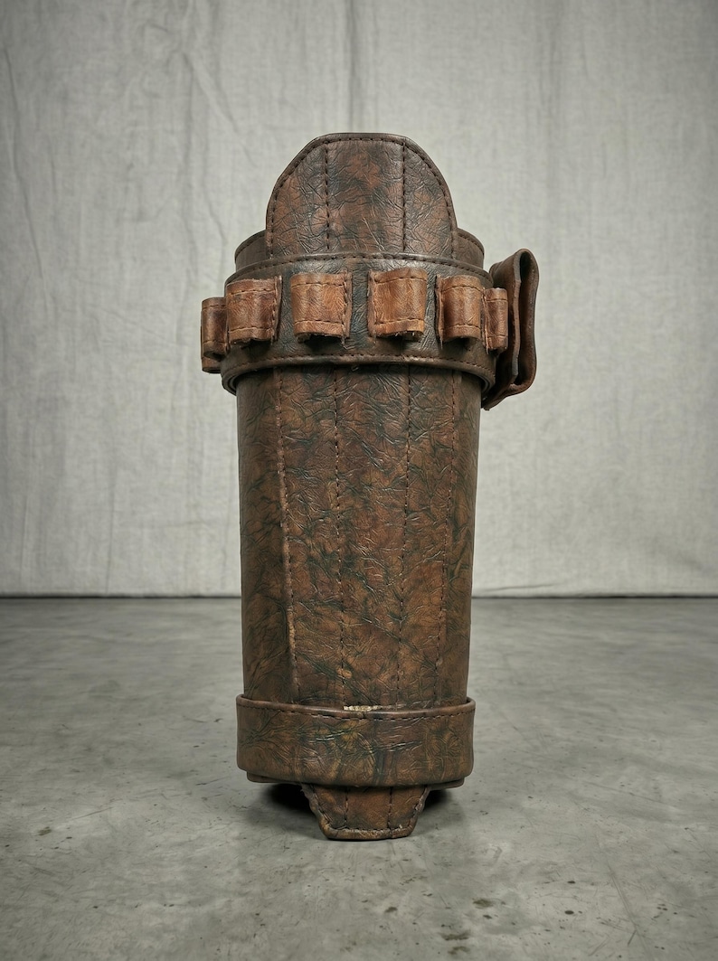 May include: A brown leather holster with a textured finish. The holster has a cylindrical shape with a series of small square pockets around the middle and a curved top. The holster is designed to hold a long, thin object.