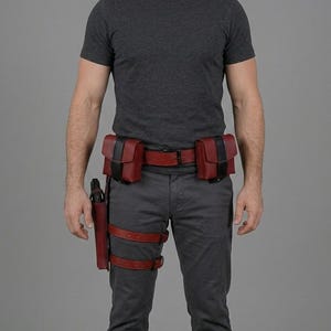 May include: A dark red leather utility belt featuring two rectangular pouches with black vertical straps, a matching holster attached to the belt, and two adjustable leg straps with black buckles, worn over a dark grey t-shirt and grey trousers.