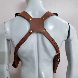 May include: A tan leather shoulder holster harness featuring adjustable straps with brass-coloured metal rivets and buckles. The harness includes two symmetrical side pouches attached to the lower ends of the straps, designed to be worn over the shoulders.
