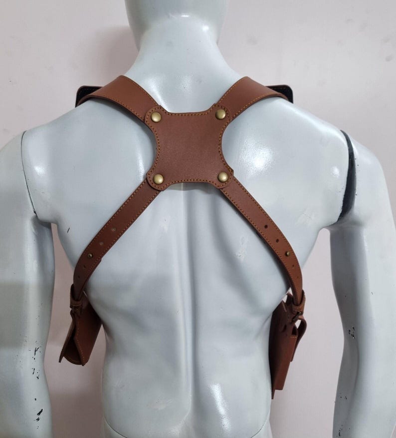 May include: A tan leather shoulder holster harness featuring adjustable straps with brass-coloured metal rivets and buckles. The harness includes two symmetrical side pouches attached to the lower ends of the straps, designed to be worn over the shoulders.