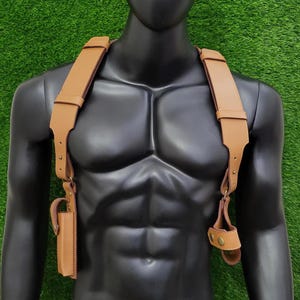 May include: A tan leather shoulder holster harness featuring wide adjustable straps, metallic hardware, and two distinct attachment points. The harness includes a vertical rectangular pouch on the right side and a circular loop fastener on the left side.