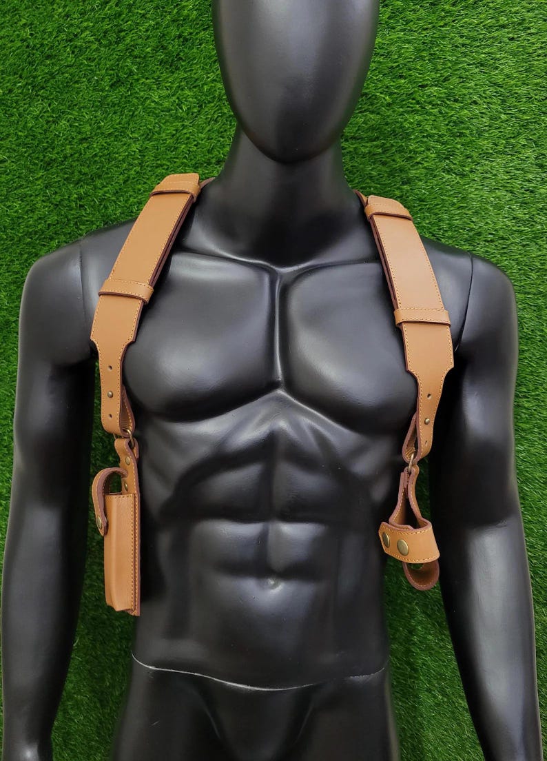 May include: A tan leather shoulder holster harness featuring wide adjustable straps, metallic hardware, and two distinct attachment points. The harness includes a vertical rectangular pouch on the right side and a circular loop fastener on the left side.