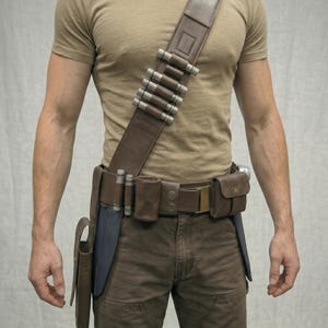 Puede incluir: Bandolera de cuero marr&oacute;n con cilindros plateados y cintur&oacute;n a juego. El cintur&oacute;n tiene bolsillos y una funda. El atuendo se completa con pantalones marrones y una camiseta beige.