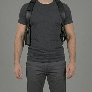 May include: A black leather shoulder holster system featuring wide adjustable straps, a rectangular pouch on the right side, and a secure loop attachment on the left side, all connected with metallic hardware over a dark grey t-shirt and trousers.