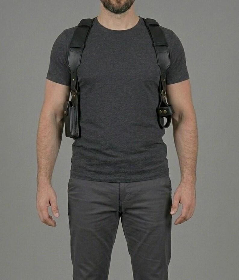 May include: A black leather shoulder holster system featuring wide adjustable straps, a rectangular pouch on the right side, and a secure loop attachment on the left side, all connected with metallic hardware over a dark grey t-shirt and trousers.