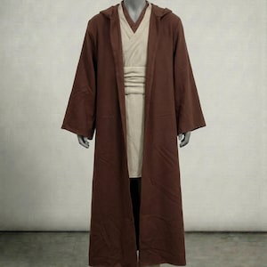 Puede incluir: Una t&uacute;nica con capucha marr&oacute;n con mangas largas, usada sobre una t&uacute;nica y faja color crema. El disfraz se exhibe en un maniqu&iacute;. El atuendo recuerda a un Jedi o personaje similar.
