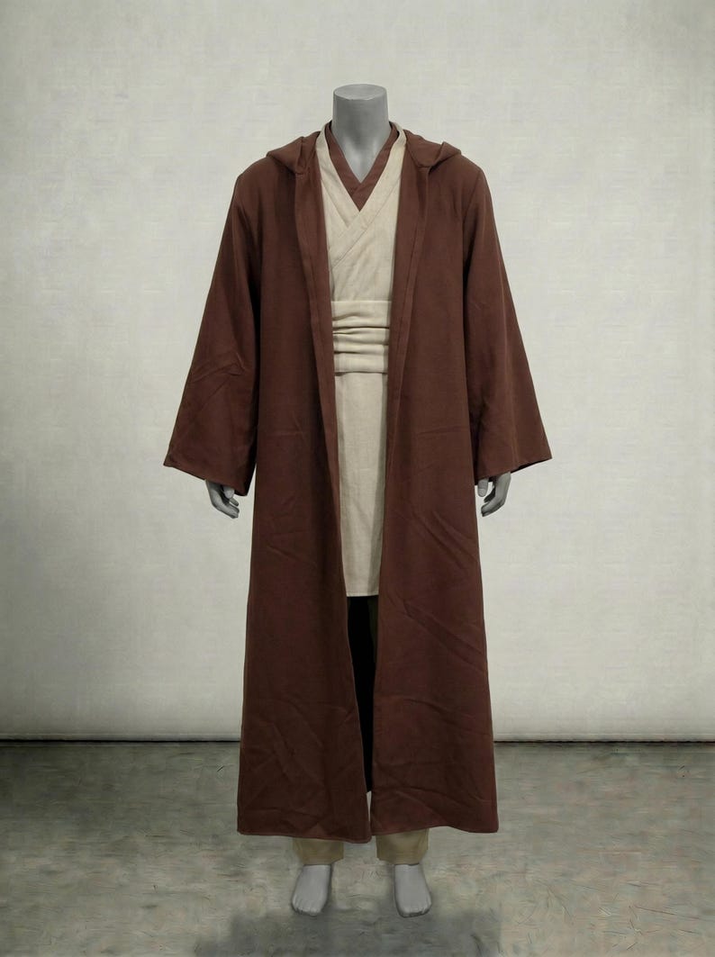 Puede incluir: Una t&uacute;nica con capucha marr&oacute;n con mangas largas, usada sobre una t&uacute;nica y faja color crema. El disfraz se exhibe en un maniqu&iacute;. El atuendo recuerda a un Jedi o personaje similar.