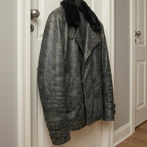 Puede incluir: Una chaqueta de cuero gris con cuello de piel negra cuelga de un gancho. La chaqueta tiene un dise&ntilde;o cruzado, varios botones y detalles de hebilla en las mangas. La chaqueta est&aacute; colgada en una puerta blanca.