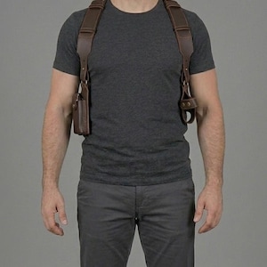 May include: A dark brown leather shoulder holster system featuring wide adjustable straps that cross over the shoulders and chest. The harness includes a rectangular pouch on the right side and a circular attachment point on the left side with metal hardware.