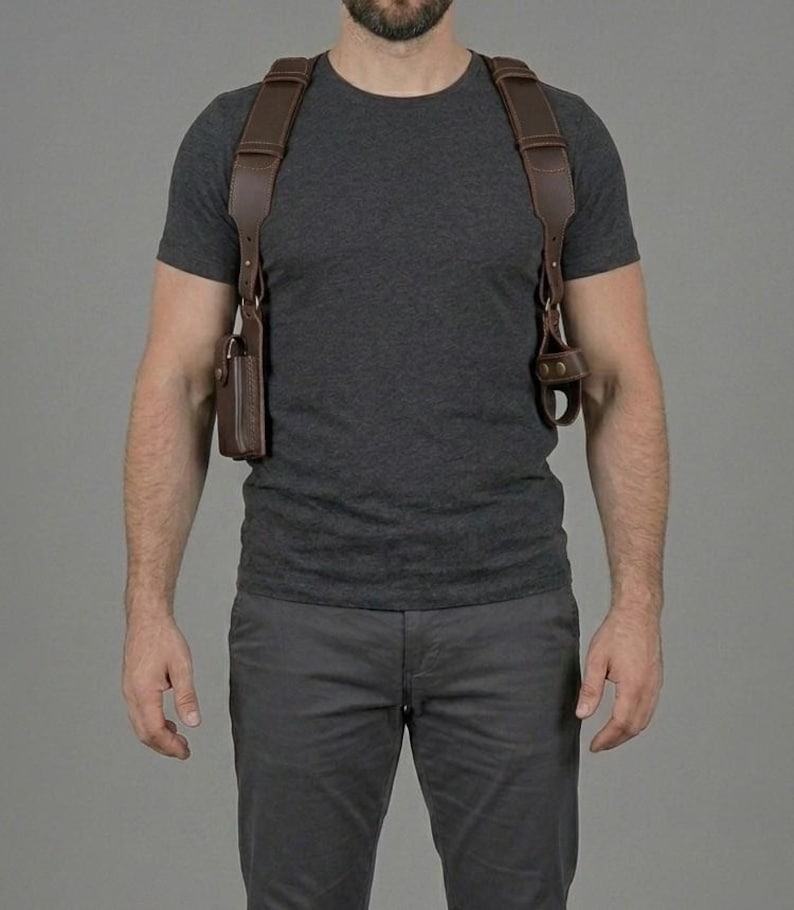 May include: A dark brown leather shoulder holster system featuring wide adjustable straps that cross over the shoulders and chest. The harness includes a rectangular pouch on the right side and a circular attachment point on the left side with metal hardware.
