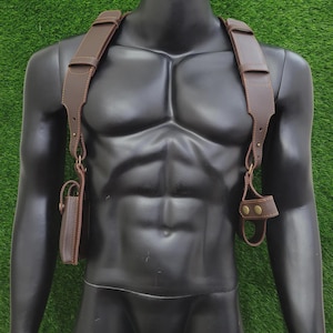 May include: A brown leather shoulder holster harness featuring wide adjustable straps, brass-toned metal hardware, and two distinct attachments. One side includes a rectangular pouch with a flap, while the other side features a circular loop holder.
