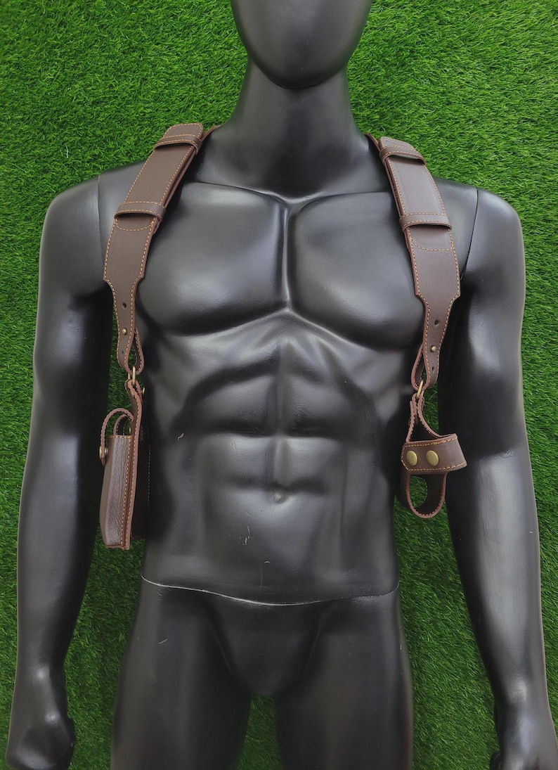 May include: A brown leather shoulder holster harness featuring wide adjustable straps, brass-toned metal hardware, and two distinct attachments. One side includes a rectangular pouch with a flap, while the other side features a circular loop holder.