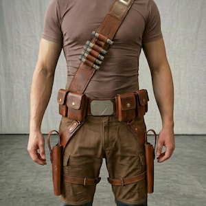 May include: A brown leather bandolier with metallic cylindrical accents worn over a brown t-shirt, paired with a matching leather utility belt featuring two side pouches, a central metallic buckle, and two vertical holsters on the thighs.