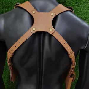 May include: A tan leather shoulder harness featuring adjustable straps with metal buckles and a central back panel secured by four brass-coloured rivets. The harness includes two side pouches attached to the lower ends of the straps.