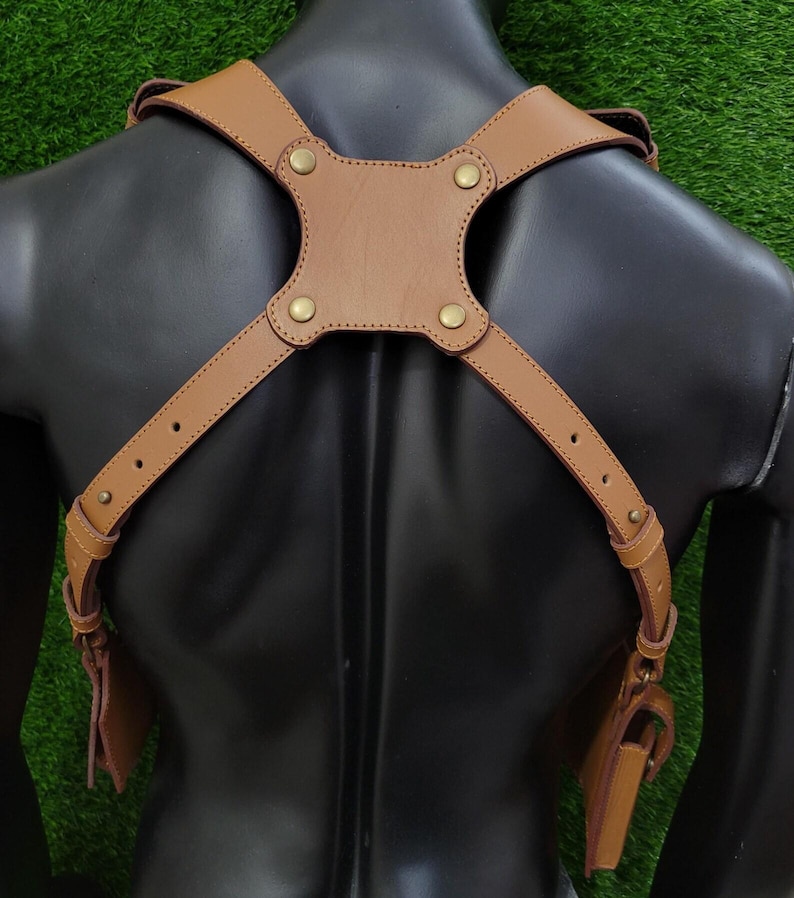 May include: A tan leather shoulder harness featuring adjustable straps with metal buckles and a central back panel secured by four brass-coloured rivets. The harness includes two side pouches attached to the lower ends of the straps.