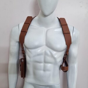 May include: A tan leather shoulder holster harness featuring wide adjustable straps that cross over the chest and back. The harness includes a rectangular pouch on the right side and a circular loop attachment on the left side with visible stitching.