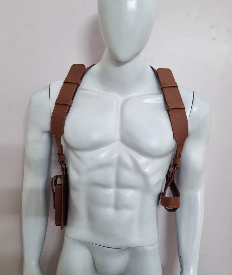 May include: A tan leather shoulder holster harness featuring wide adjustable straps that cross over the chest and back. The harness includes a rectangular pouch on the right side and a circular loop attachment on the left side with visible stitching.