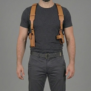 May include: A tan leather shoulder holster system featuring wide shoulder straps and two side-mounted holsters with snap closures. The harness is worn over a dark grey t-shirt and is paired with dark grey trousers and a matching grey tactical belt.