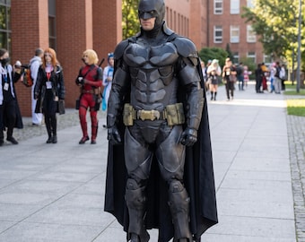 Könnte beinhalten: Ein Batman-Ganzkörperkostüm bestehend aus einer schwarzen, geformten Rüstung mit spitzer Maske, einem langen, fließenden schwarzen Umhang und einem Gürtel mit goldenen und olivgrünen Taschen. Der Anzug zeigt segmentierte Brust-, Arm- und Beinplatten.