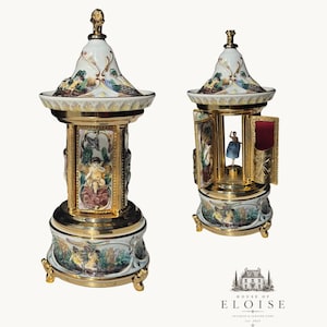 May include: A gold and white porcelain music box featuring a conical lid, a central rotating ballerina figure, and hinged panels decorated with colorful painted cherubs and landscape scenes. The base is supported by three ornate gold feet.