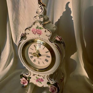 Puede incluir: Un reloj de sobremesa de cerámica blanca con bordes dorados, motivos florales rosas pintados a mano y rosas esculpidas en la base. La esfera tiene números romanos negros, manecillas doradas y el nombre de la marca Mercedes en texto negro.