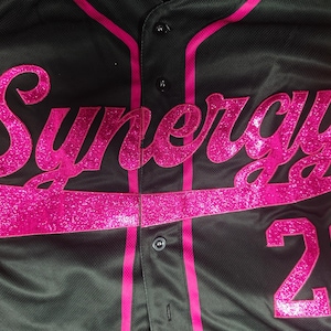 Pode incluir: Uma camisa de beisebol preta com bot&otilde;es, apresentando a palavra Synergy em letras cursivas grandes e rosa brilhante no peito. A camisa inclui debrum rosa ao longo da carcela frontal e o n&uacute;mero 26 em rosa brilhante na parte inferior direita.