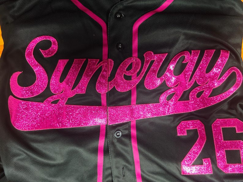 Pode incluir: Uma camisa de beisebol preta com bot&otilde;es, apresentando a palavra Synergy em letras cursivas grandes e rosa brilhante no peito. A camisa inclui debrum rosa ao longo da carcela frontal e o n&uacute;mero 26 em rosa brilhante na parte inferior direita.