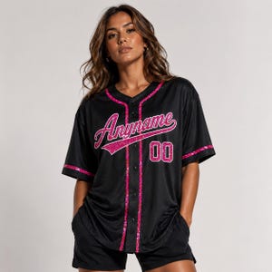 Pode incluir: Uma camisola de basebol preta com fecho de bot&otilde;es, mangas curtas e acabamentos em glitter rosa brilhante ao longo da gola, da carcela e dos punhos. A parte da frente apresenta a palavra Anyname em letra cursiva rosa e o n&uacute;mero 00 em rosa.