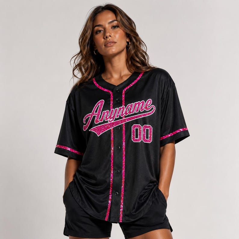 Pode incluir: Uma camisola de basebol preta com fecho de bot&otilde;es, mangas curtas e acabamentos em glitter rosa brilhante ao longo da gola, da carcela e dos punhos. A parte da frente apresenta a palavra Anyname em letra cursiva rosa e o n&uacute;mero 00 em rosa.