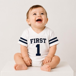 May include: A white short-sleeved baby bodysuit with a V-neckline, featuring dark blue stripes on the sleeves and the text FIRST 1 printed in dark blue on the chest. The garment is designed with a sporty aesthetic, resembling a football jersey.