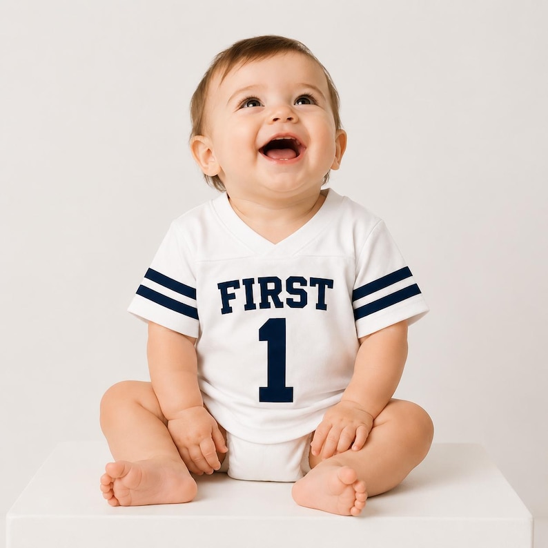 May include: A white short-sleeved baby bodysuit with a V-neckline, featuring dark blue stripes on the sleeves and the text FIRST 1 printed in dark blue on the chest. The garment is designed with a sporty aesthetic, resembling a football jersey.