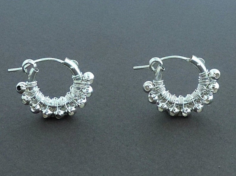 May include: A pair of silver hoop earrings with small silver beads attached to the hoop. The earrings are made of wire and have a delicate, handcrafted look.