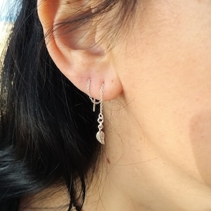 May include: A silver chain earring with a small leaf charm hanging from it. The chain is attached to a small hoop earring. The earring is worn in the upper lobe of the ear.