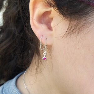 May include: Silver chain earrings featuring a small, dangling red gemstone. The earrings connect two ear piercings with a delicate chain, and the gemstone is set in a silver bezel. The earrings are shown on a person's ear.