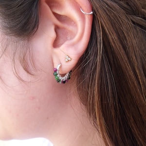 Op de afbeelding: Een close-up van iemands oor met een zilveren oorring met groene en roze edelstenen. De oorring zit in de onderste oorlel. Er zit een klein gouden driehoekig oorbelletje in de bovenste oorlel.