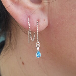 May include: A silver chain earring with a teardrop-shaped blue gemstone dangling from the end. The chain is threaded through two piercings in the ear.