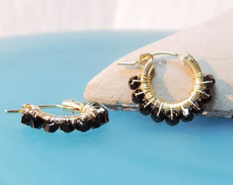 Puede incluir: Aretes de aro dorados con detalles de piedras preciosas negras. Los aretes están hechos con alambre envuelto alrededor de las piedras preciosas.